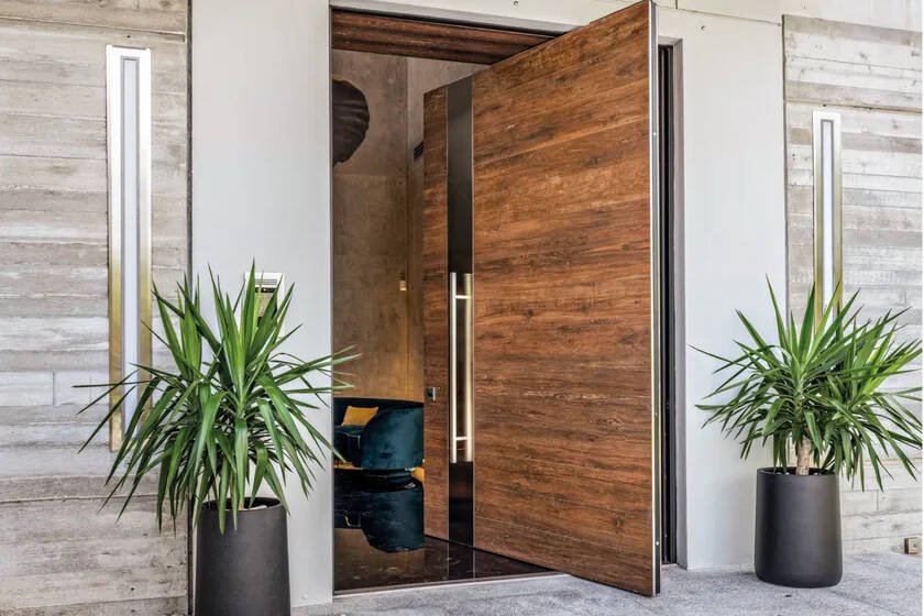 Open wood-finished pivot entrance door with a long metal pull handle between two planters